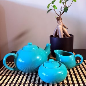 Peut inclure: Un service à thé en céramique turquoise vif composé d'une théière ronde avec couvercle, d'un sucrier assorti avec couvercle et d'un pot à lait. Les pièces présentent une finition lisse et brillante et sont disposées sur une surface plane.