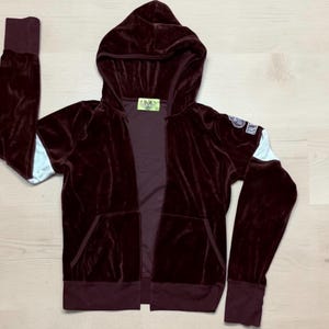 Puede incluir: Una sudadera con capucha y cremallera de terciopelo color burdeos oscuro, con capucha, mangas largas con paneles azul claro, puños acanalados y dos bolsillos delanteros. Un pequeño parche rectangular con texto blanco es visible en la manga izquierda.