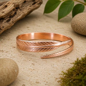May include: A copper-colored metal cuff bracelet shaped like a feather. The bracelet features a wrap-around design with detailed etched lines mimicking the structure of a feather, including a central vein and fine barbs along the entire length.