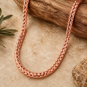 Pode incluir: Um colar de corrente em ouro rosa com um design tecido. O colar é exibido em um pedaço de madeira e uma superfície de pedra. A corrente é de uma cor metálica quente, e o padrão tecido dá-lhe uma aparência texturizada.