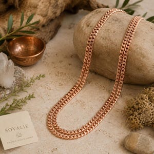 May include: A copper-colored curb chain necklace is draped over a stone. The necklace is made of interlocking links. A small copper bowl, quartz crystals, and a card with the text "SOVALIE" are in the background. The card also reads "BALANCE. BEAUTY. ENERGY."