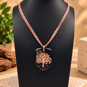 May include: A copper-colored curb chain necklace featuring a shield-shaped pendant. The pendant has a black surface with an embedded metallic copper-colored tree design with intricate, swirling branches and a solid trunk centered on the front.