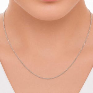 Puede incluir: Un delicado collar de cadena en tono plateado que presenta una serie de pequeños eslabones ovalados uniformes. El metal tiene un acabado pulido y reflectante que capta la luz, creando un brillo sutil a lo largo de la cadena fina y ligera.