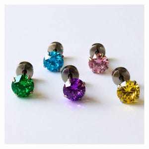 Puede incluir: Cinco pendientes de botón con piedras preciosas talladas en forma redonda en colores vibrantes que incluyen verde, azul brillante, rosa suave, morado intenso y amarillo. Cada piedra está engastada en una montura metálica oscura.