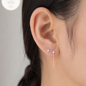Puede incluir: Primer plano de una oreja adornada con dos pares de pendientes de plata. Un par presenta tachuelas en forma de estrella, mientras que el otro tiene una tachuela de estrella con una barra colgante. Los pendientes son minimalistas y elegantes.