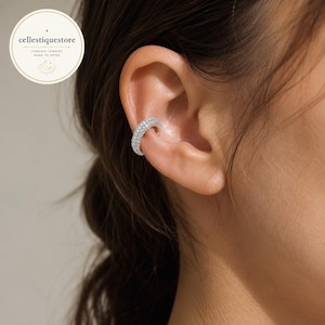Puede incluir: Un ear cuff de tono plateado con una banda gruesa y curva incrustada con numerosas piedras pequeñas, claras y brillantes. La pieza de joyería está colocada en la zona del cartílago de la oreja, mostrando un engaste denso de estilo pavé.