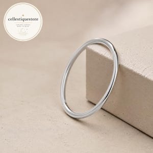 Puede incluir: Un anillo fino de metal plateado pulido con una banda lisa y redondeada. El anillo es circular y presenta una superficie metálica reflectante. Un logotipo circular en la esquina superior izquierda contiene el texto cellestiquestore, timeless jewelry, made to shine.