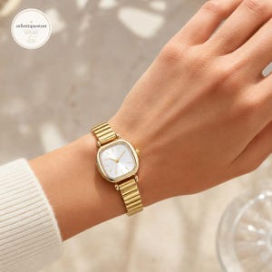 May include: A gold-toned wristwatch with a square face and a white dial. The watch has a gold band and is worn on a wrist. The watch face has gold hands and markers. The watch is a classic and elegant accessory.