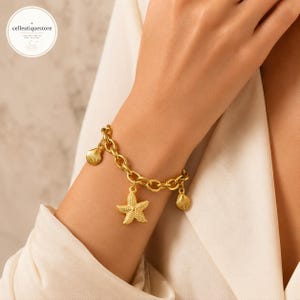 Puede incluir: Una pulsera de eslabones dorados con tres dijes colgantes: una estrella de mar texturizada en el centro y dos dijes de conchas marinas más pequeños a los lados. El metal tiene un acabado pulido y los eslabones son uniformes.