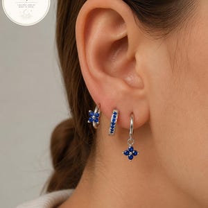Puede incluir: Tres pendientes de aro plateados en una sola oreja. El primero es un aro pequeño con un dije de flor de cuatro pétalos de piedra azul. El segundo es un aro fino incrustado con pequeñas piedras azules. El tercero es un aro pequeño con flor.