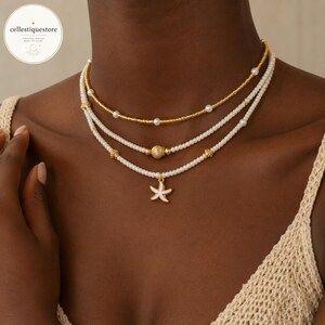 Puede incluir: Un collar de tres capas que presenta una cadena superior de cuentas doradas con perlas blancas, una hebra central de perlas blancas con una cuenta de concha dorada y una hebra inferior de perlas blancas con un dije de estrella de mar blanca y dorada.