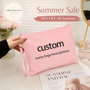 Puede incluir: Un estuche rectangular de color rosa claro con cremallera y una correa de muñeca. La parte frontal del estuche presenta el texto impreso en negro "custom" seguido de "name/logo/text/picture" en un tamaño de fuente menor.