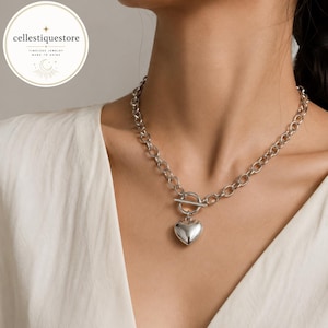 Puede incluir: Un collar de cadena en tono plateado con un cierre de barra y argolla, y un colgante liso en forma de corazón abombado. El collar se lleva alrededor del cuello, destacando los eslabones metálicos y el dije central de corazón.