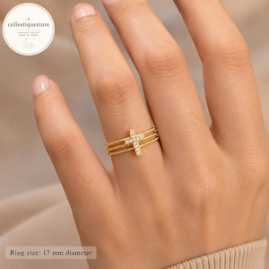 Puede incluir: Un anillo de tono dorado que presenta tres bandas delgadas y con cuentas, conectadas por un elemento de cruz vertical central. La cruz está adornada con pequeñas piedras claras y brillantes. El anillo tiene un diámetro indicado de 17 mm.