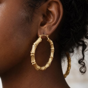 Puede incluir: Pendientes de aro dorados con diseño de bambú. Los pendientes presentan un diseño texturizado que imita el aspecto de los tallos de bambú. Los aros se llevan en la oreja, con un segundo pendiente visible en el fondo.