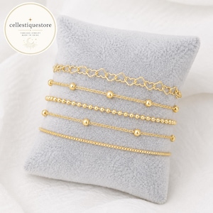 Puede incluir: Un conjunto de cinco pulseras doradas sobre un cojín gris suave. La colección incluye una cadena de eslabones en forma de corazón, dos cadenas de cuentas de distintos tamaños, una cadena fina con cuentas espaciadas y una pulsera de cadena cuadrada.