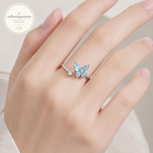 Puede incluir: Anillo de mariposa plateado con detalles de piedras preciosas azul claro. El anillo presenta un diseño abierto con una pequeña gema transparente en un lado y una mariposa con cuatro piedras azules en el otro. El anillo está en un dedo.