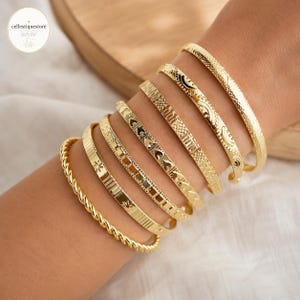 Puede incluir: Una colección de pulseras doradas con varios diseños, incluyendo estilos texturizados, estampados y trenzados. Las pulseras se muestran en una muñeca, mostrando sus diferentes texturas y formas. La joyería es probablemente de metal.