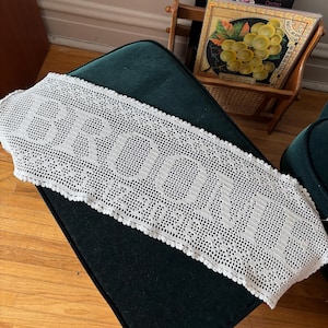 May include: A rectangular off-white crochet runner featuring an intricate openwork pattern. The word BROOM is prominently displayed in the center using a solid stitch technique, with a decorative scalloped edge bordering the entire piece.