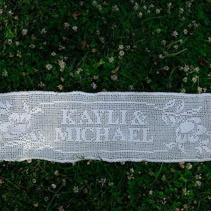 May include: A rectangular white crochet runner featuring a delicate floral pattern on both ends and the text KAYLI & MICHAEL centered in the middle. The piece is crafted with a fine grid-like stitch, creating a detailed and intricate design.