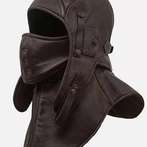 Può includere: Un copricapo in pelle marrone scuro caratterizzato da un design a cappuccio a copertura totale con maschera facciale integrata. La maschera presenta piccoli occhielli metallici di ventilazione, mentre il cappuccio ha cinturini laterali regolabili.