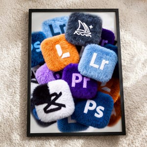 May include: A framed print featuring a collection of square, plush, faux-fur icons in various colors including orange, light blue, purple, and white. Each icon displays a white or black embroidered software logo, such as Lr, Pr, Ps, and a sailboat.
