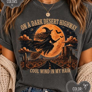 Puede incluir: Una camiseta gris carbón con un gráfico de una bruja volando en una escoba frente a una gran luna naranja. El texto dice ON A DARK DESERT HIGHWAY y COOL WIND IN MY HAIR. La escena incluye un paisaje desértico con cactus y una carretera.