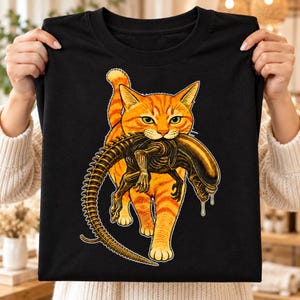 May include: A black t-shirt featuring a graphic illustration of an orange tabby cat walking forward while carrying a dark, skeletal, alien-like creature in its mouth. The cat has green eyes and the creature has a long, segmented tail.