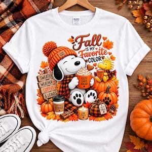 Może przedstawiać: Biały t-shirt z grafiką psa z kreskówki w pomarańczowej czapce, trzymającego kubek z napisem Cozy Days Ahead, oraz tekstami Fall Is My Favorite Color, Sweater Weather, Coffee & Leaves i Stay Cozy.