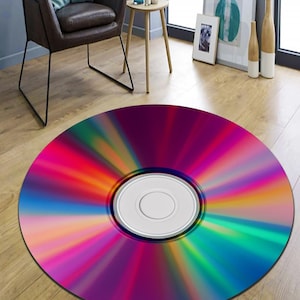 May include: A round rug with a vibrant, multicolored design resembling a CD. The rug features a central white circle and radiating bands of color, including purple, red, orange, yellow, green, and blue. It is placed on a wooden floor.