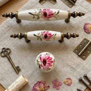 May include: Decorative cabinet hardware set featuring two white ceramic handles and a round knob, each adorned with a pink floral design. Includes brass hinges, screws, and a vintage key, ideal for home decor projects.