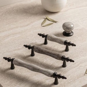 May include: Three cabinet pulls and one matching round cabinet knob. Each piece features a smooth, light gray marble-like center section with dark brown, metallic-finished end caps and mounting posts, showcasing a classic, elongated design.