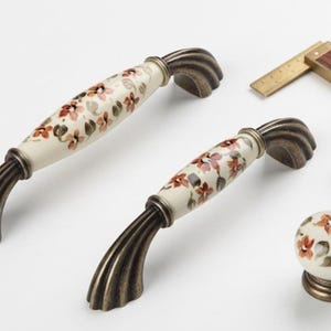 Könnte beinhalten: Zwei gewölbte Schrankgriffe und ein runder Schrankknopf mit einem cremefarbenen Keramikmittelteil mit aufgemaltem orange-grünem Blumenmuster, abgerundet durch Endstücke aus antik bronzefarbenem Metall. Ein kleiner Metallwinkel ist sichtbar.