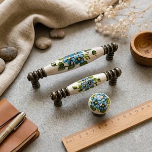 Può includere: Due maniglie per cassetti in ceramica e un pomello rotondo in ceramica con fiori blu e foglie verdi dipinti a mano su sfondo bianco. La ferramenta presenta estremità in metallo color bronzo antico con dettagli a coste. Un righello in legno è visibile.