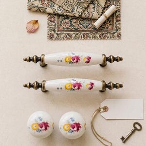 Puede incluir: Tiradores y pomos de cerámica blanca con diseños florales. Los tiradores tienen extremos de color bronce. Se ven dos pomos y dos tiradores, junto con una llave vintage y una etiqueta en blanco. Una pequeña alfombra y un papel enrollado están en el fondo.