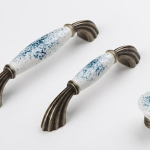 Puede incluir: Dos tiradores de armario arqueados y un pomo redondo con centro de cerámica blanca y patrón de motas azules. Los herrajes incluyen extremos de metal en tono bronce antiguo con un diseño acanalado en las bases de montaje.