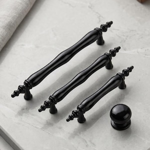 May include: A set of four black cabinet hardware pieces. Three are elongated handles with decorative ends, and one is a round knob. The hardware is displayed on a light gray marble surface.
