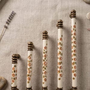 May include: Five cylindrical cabinet pulls of varying lengths, each featuring a cream-colored ceramic center decorated with a repeating pattern of small orange flowers and green leaves, finished with ribbed antique brass metal end caps.