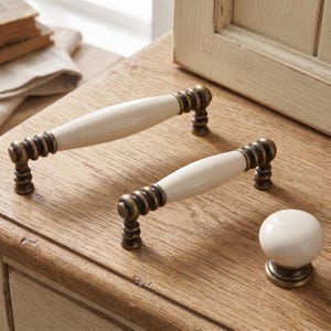 May include: Two cabinet pulls and one round cabinet knob. Each pull features a smooth, cream-colored ceramic center handle with antique brass-finished metal ends that have a ribbed, tiered design. The knob is a matching cream-colored ceramic sphere.