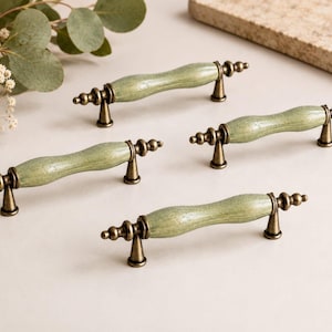 May include: Four sage green cabinet pulls with a wavy design and antique brass finish. The pulls are arranged on a light surface, showcasing their decorative details and hardware.