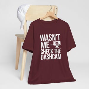 Puede incluir: Una camiseta granate con el texto blanco "WASNT ME - CHECK THE DASHCAM". La camiseta está colgada de una percha de madera. Una gorra de béisbol blanca descansa sobre el respaldo de una silla de madera.