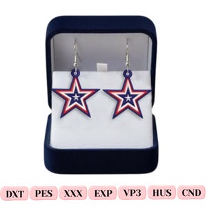 May include: A pair of star-shaped dangle earrings featuring a layered design in red, white, and blue, suspended from silver-toned metal hooks. The earrings are displayed inside a dark blue velvet jewelry box with a white interior lining.