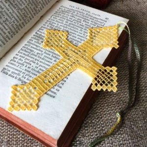 May include: A yellow lace cross bookmark with a delicate grid pattern and pointed ends, featuring a thin matching thread attached to the top, resting on the open pages of an antique book with printed text in a dark serif font.