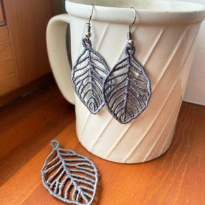May include: A pair of grey leaf-shaped earrings made of intricate thread work, featuring silver-toned metal hooks. A single matching grey leaf-shaped thread ornament is positioned separately in the foreground, resting near a white ceramic mug.