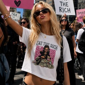Può includere: Una t-shirt bianca corta con una stampa grafica di una bambola seduta su un mucchio di soldi con un cagnolino, accompagnata dal testo EAT THE RICH. La maglietta ha maniche corte e scollo rotondo, indossata con una cintura nera con borchie e borsa.