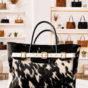 Op de afbeelding: Een grote tote bag met een zwart-wit koeienhuidpatroon met een textuur van bont. De tas heeft twee zwarte lederen handvatten en een horizontale witte lederen riem aan de voorzijde, vastgezet met drie goudkleurige metalen gespen.