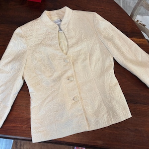 May include: A pale yellow blazer featuring a tonal swirling paisley pattern, a mandarin collar, and a front opening with three matching fabric-covered buttons. The jacket has three-quarter length sleeves and a structured, tailored fit.