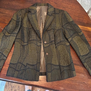 May include: An olive green blazer featuring a patchwork design with visible dark stitching along the seams. The jacket has a notched lapel, long sleeves, and two brown buttons on the front closure. The interior is lined with a solid tan fabric.