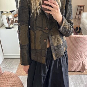 May include: A long-sleeve blazer in shades of olive green and dark brown featuring a textured, patchwork-style fabric with visible dark stitching. The garment has a notched collar, two front buttons, and a structured fit, paired with a black skirt.