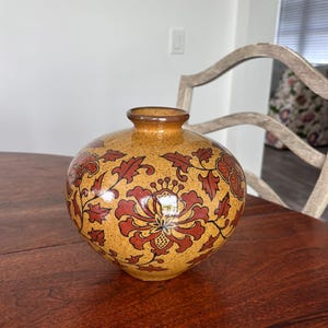 May include: A round ceramic vase with a speckled mustard yellow glaze, featuring a repeating pattern of dark red floral motifs and leafy vines. The vase has a short, narrow neck with a smooth, rounded rim and a glossy finish throughout.